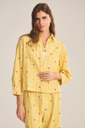 A woman wears the ASTREN SHIRT by Velvet by Graham & Spencer, a yellow checked embroidered shirt with small red flower patterns, paired with matching pants, standing against a plain background.