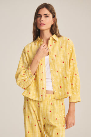 A woman wears the ASTREN SHIRT by Velvet by Graham & Spencer, a yellow striped long-sleeve embroidered top with red floral accents, paired with matching pants over a white shirt.