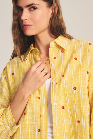 A woman wears the ASTREN SHIRT by Velvet by Graham & Spencer—a yellow cropped button-up with white stripes and red floral embroidery—over a white top, holding her necklace with one hand.