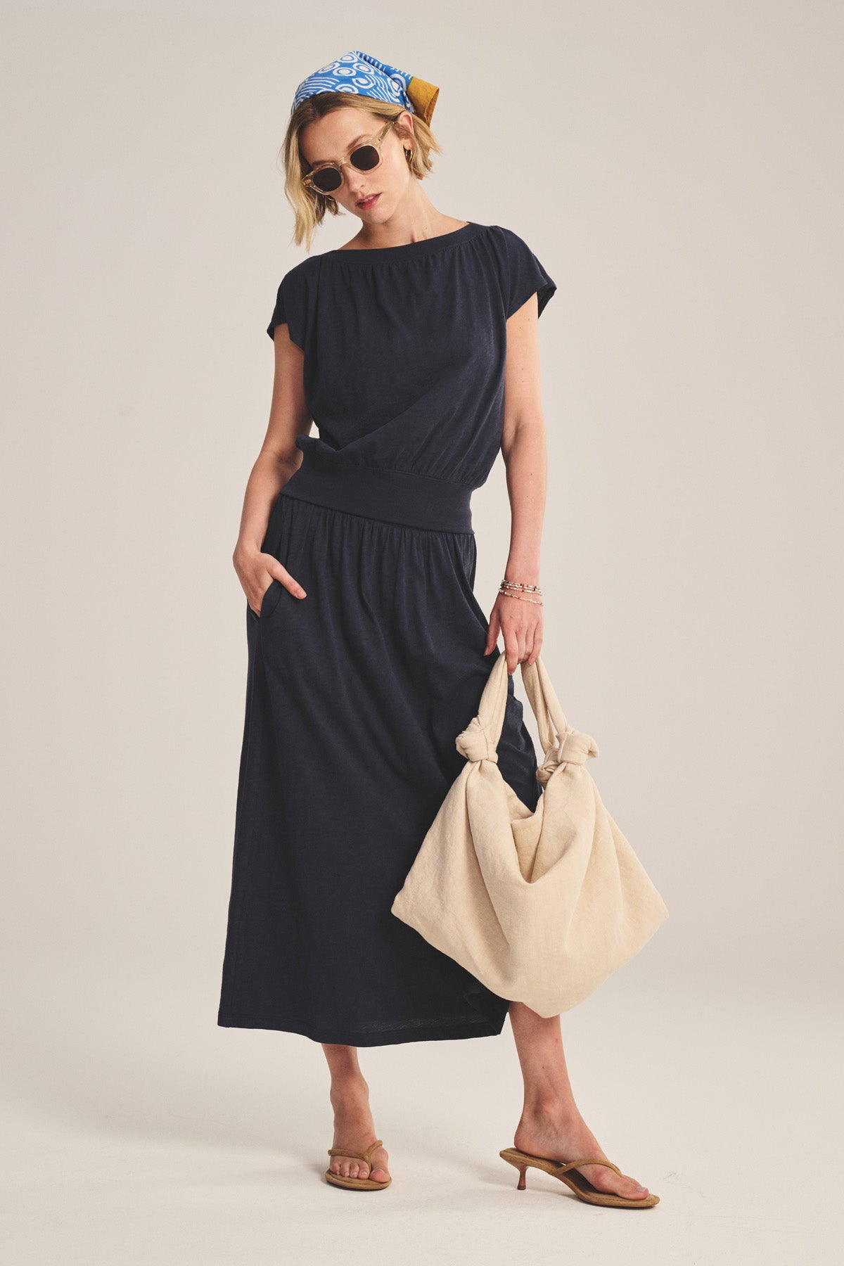 A woman in the AUDEN TEE by Velvet by Graham & Spencer, styled with tan sandals, sunglasses, a blue patterned headscarf, and a large beige knotted bag, stands against a plain background.