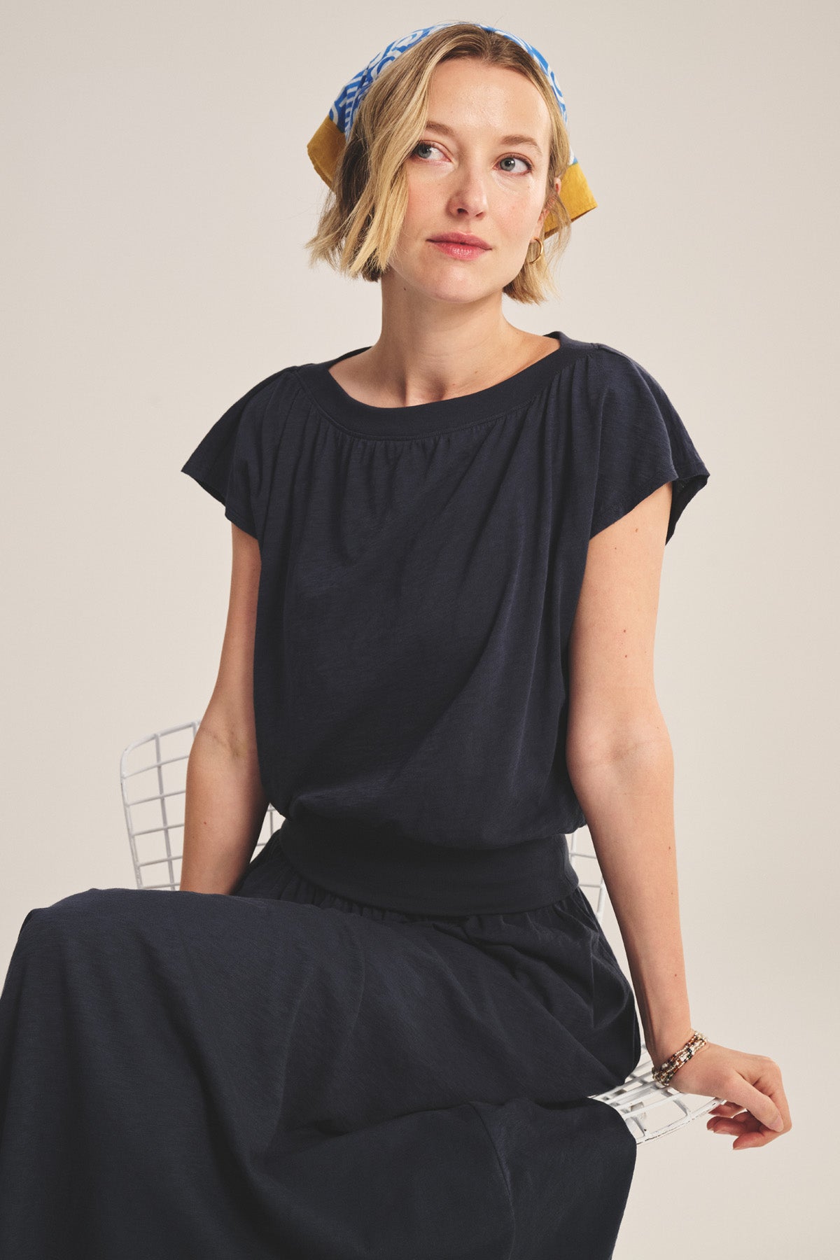 A woman with short blonde hair wears the navy AUDEN TEE by Velvet by Graham & Spencer and a patterned headscarf, seated on a white chair against a neutral background.