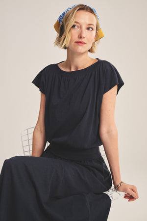 A woman with short blonde hair wears the navy AUDEN TEE by Velvet by Graham & Spencer and a patterned headscarf, seated on a white chair against a neutral background.