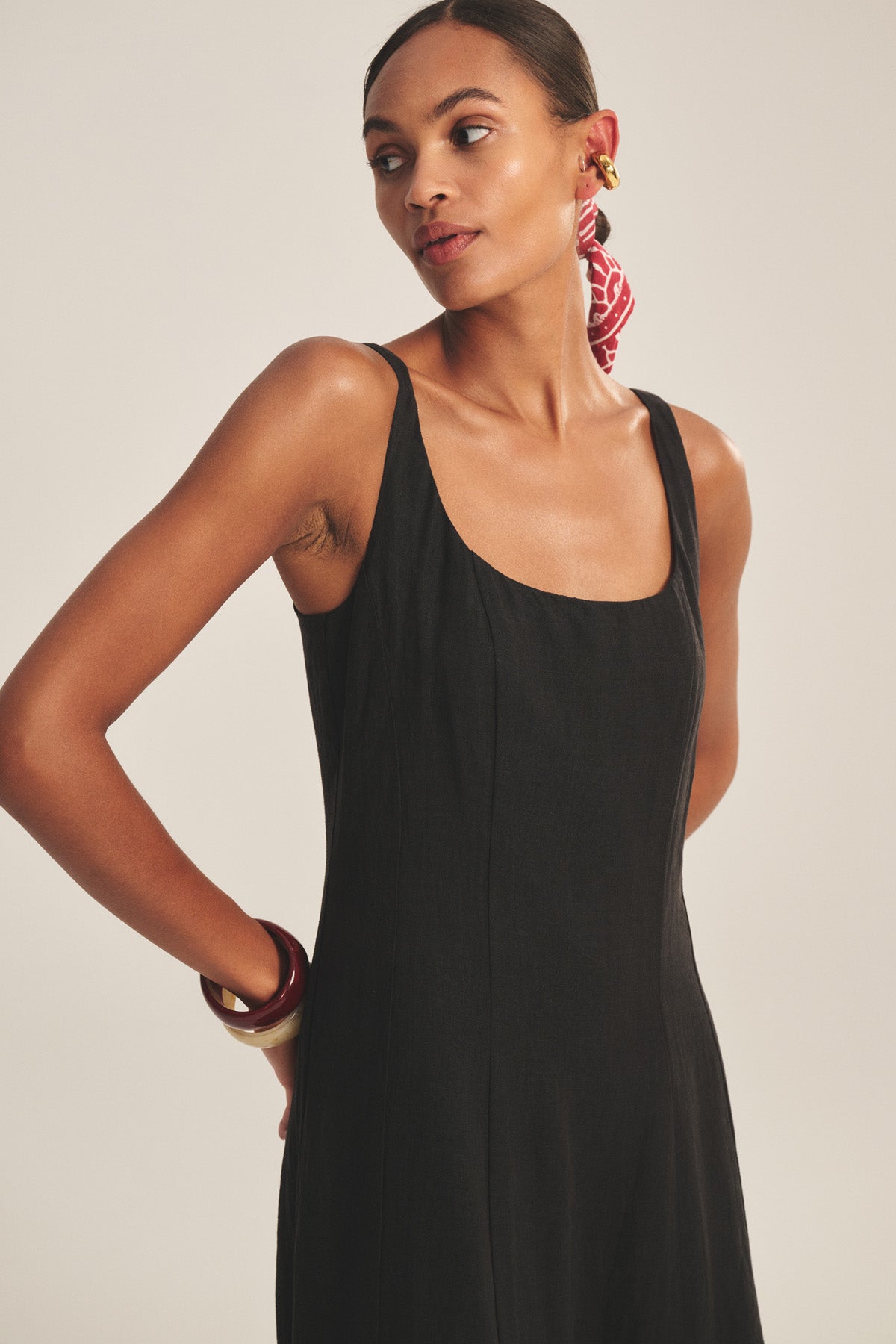 A woman in the AUSTEN LINEN DRESS by Velvet by Graham & Spencer, a sleeveless black midi with a tailored fit, looks to the side. She accessorizes with a red patterned scarf in her hair, gold hoop earrings, and chunky bracelets.-41080161009857