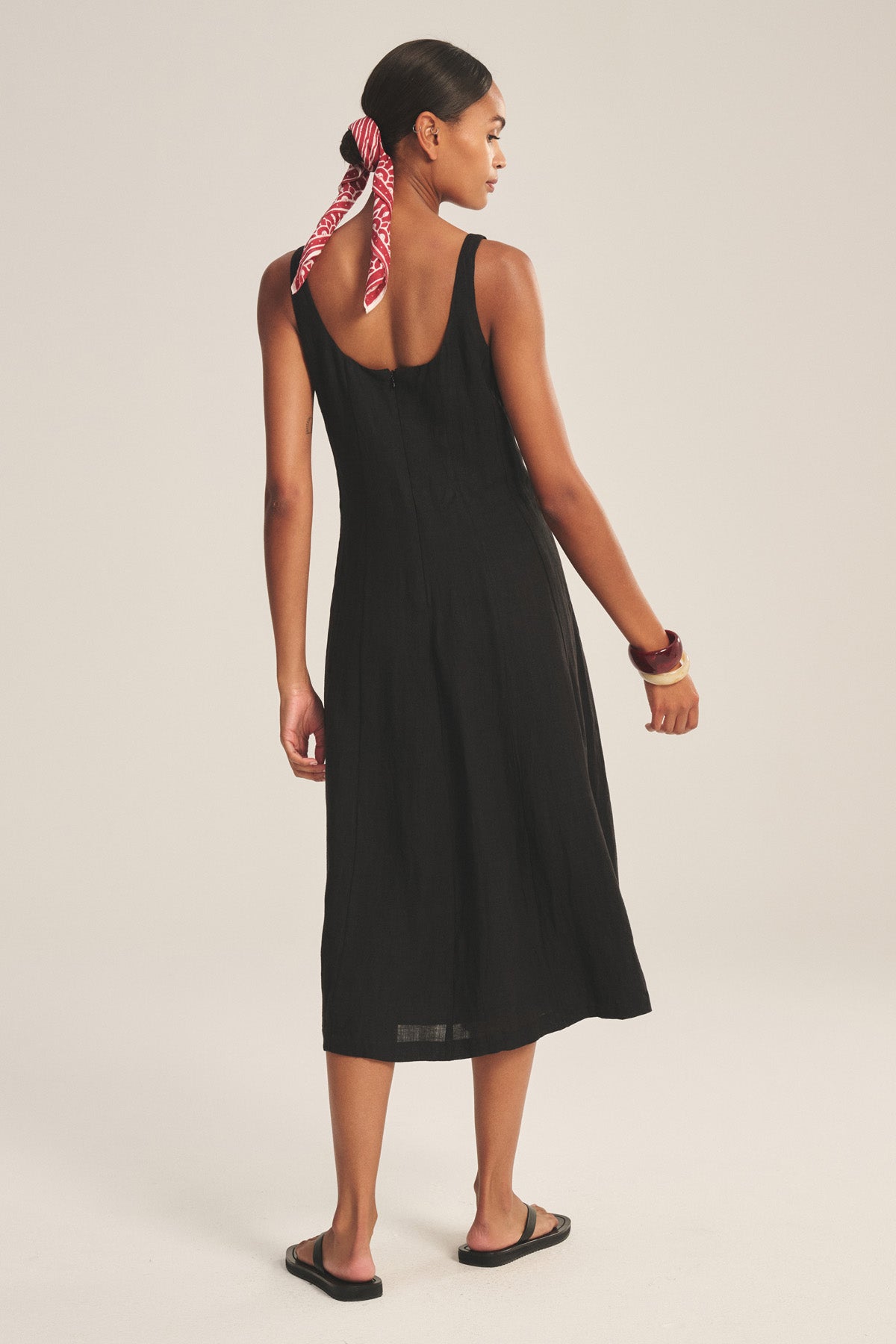 A woman faces away wearing Velvet by Graham & Spencer's AUSTEN LINEN DRESS in black, paired with black sandals, a red patterned hair scarf, and a bracelet on her right wrist.-41080161075393