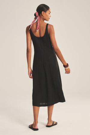 A woman faces away wearing Velvet by Graham & Spencer's AUSTEN LINEN DRESS in black, paired with black sandals, a red patterned hair scarf, and a bracelet on her right wrist.