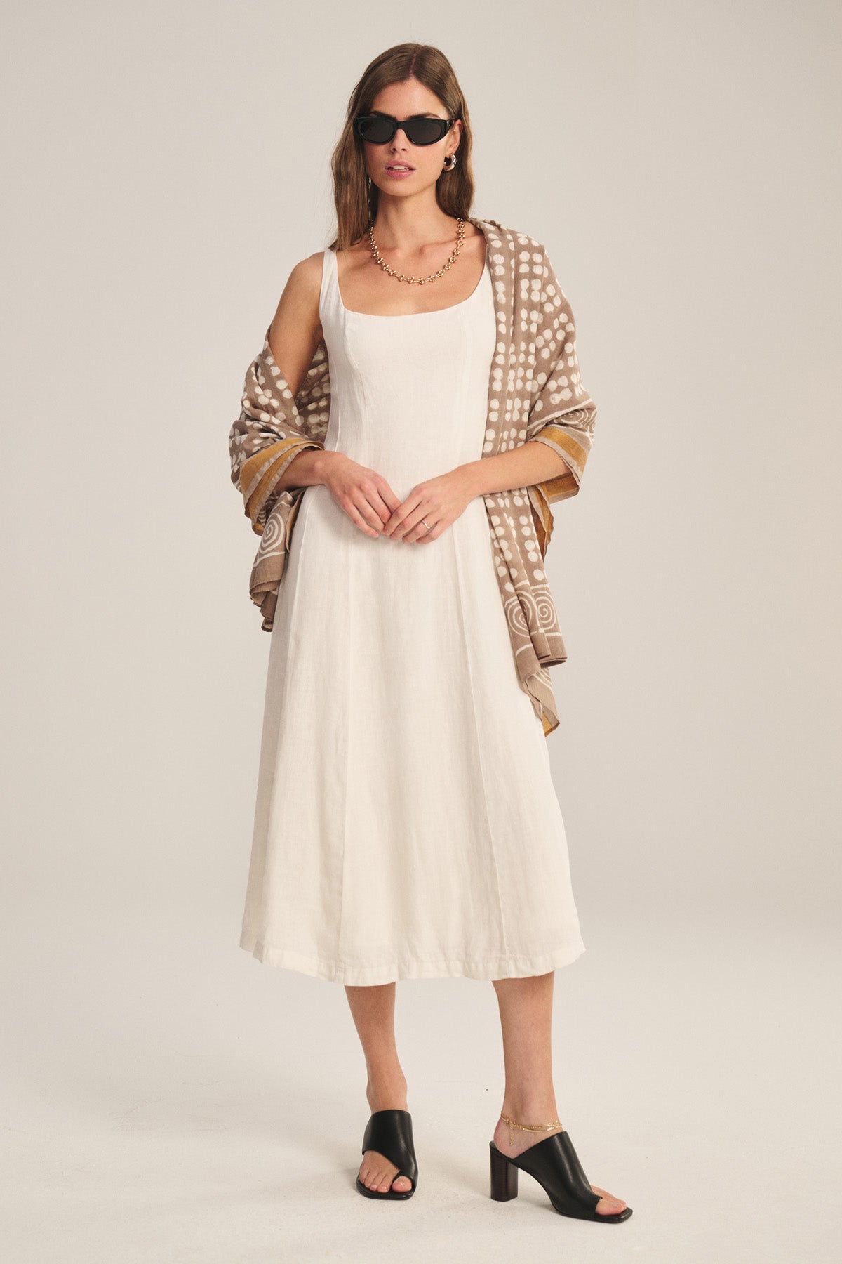 A woman wears the AUSTEN LINEN DRESS by Velvet by Graham & Spencer, styled with black heeled sandals, dark sunglasses, and a patterned brown shawl, standing against a plain light background.