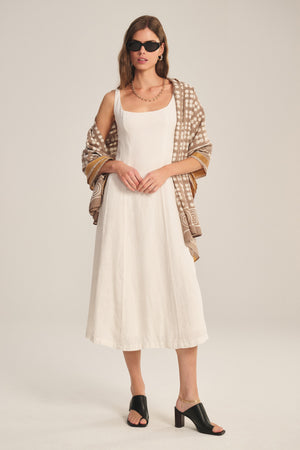 A woman wears the AUSTEN LINEN DRESS by Velvet by Graham & Spencer, styled with black heeled sandals, dark sunglasses, and a patterned brown shawl, standing against a plain light background.