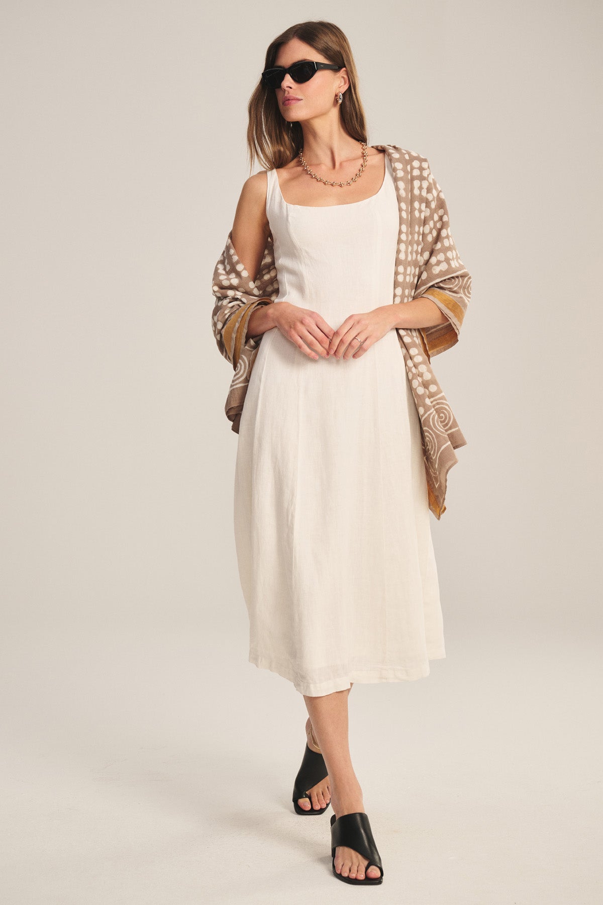 A woman stands against a plain background wearing the AUSTEN LINEN DRESS by Velvet by Graham & Spencer, a sleeveless white midi dress with a tailored silhouette, paired with a patterned brown shawl, black slide sandals, and sunglasses.-41080161173697