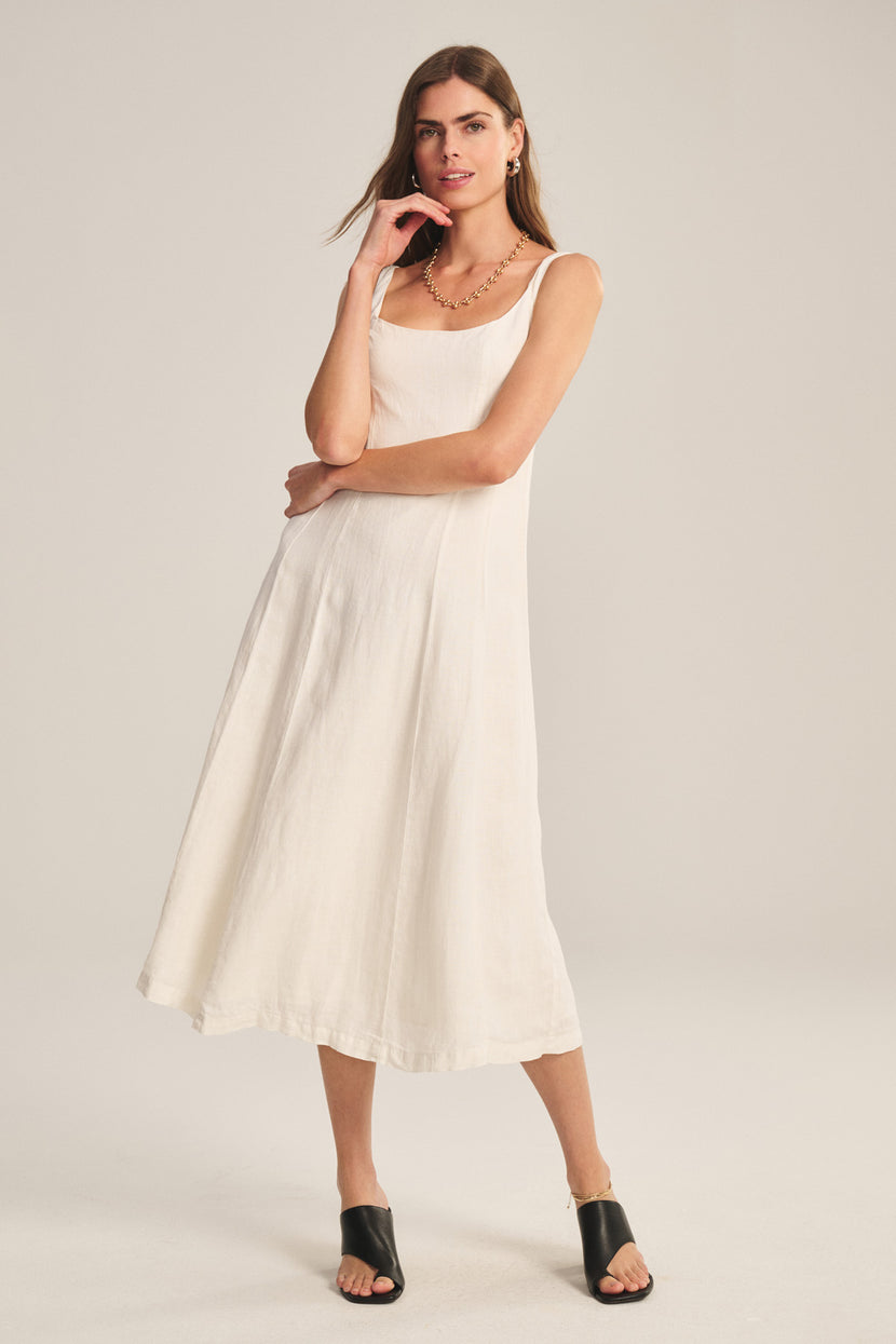 A woman stands against a plain background wearing the AUSTEN LINEN DRESS by Velvet by Graham & Spencer—a sleeveless, breathable linen midi with a tailored silhouette—paired with black open-toe sandals.
