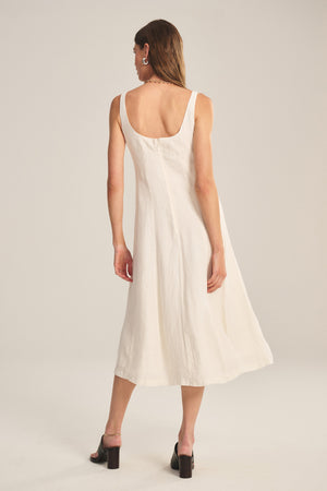 A woman stands facing away in the AUSTEN LINEN DRESS by Velvet by Graham & Spencer—an off-white, sleeveless, tailored midi with a scoop back—paired with black heeled sandals against a plain background.