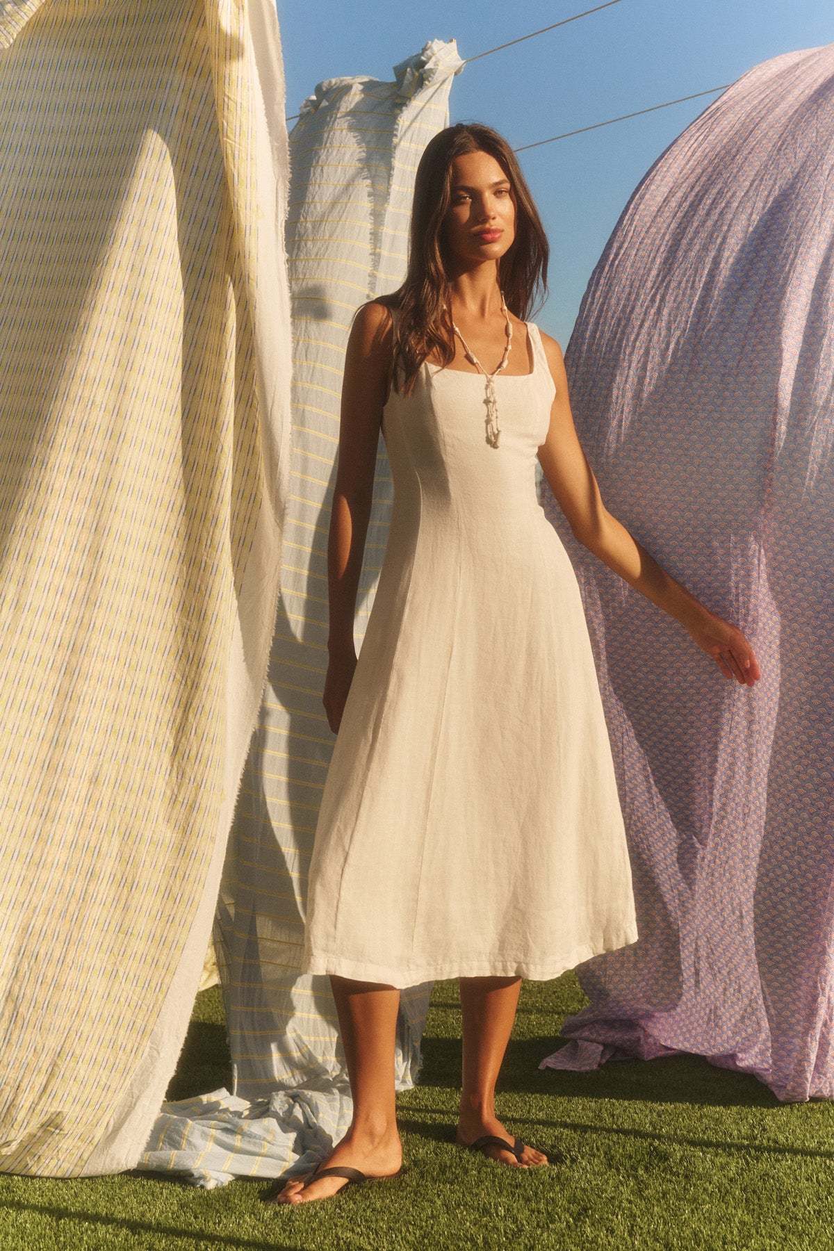 Wearing the AUSTEN LINEN DRESS by Velvet by Graham & Spencer, a woman in sandals stands on grass beside sunlit, patterned fabrics.-41080161435841