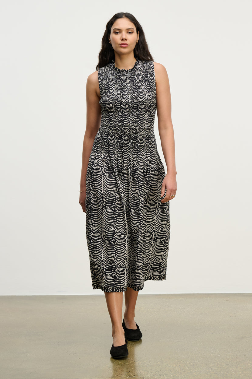 A woman poses against a plain background in the KATHERINE DRESS by Velvet by Graham & Spencer—a sleeveless, black-and-white patterned cotton midi with a smocked bodice—paired with black flats.