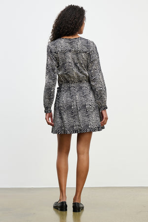 A woman faces away, wearing the MADISON DRESS by Velvet by Graham & Spencer—a black and white zebra-print cotton dress with long sleeves and subtle ruffles—paired with black shoes, set against a plain light background.