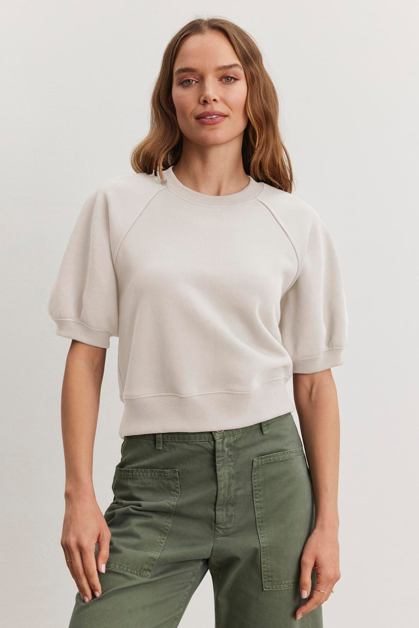 Someone wearing the BERNADETTE SWEATSHIRT by Velvet by Graham & Spencer, a light-colored cropped short-sleeve design in soft cotton fleece, and green pants stands against a plain background.