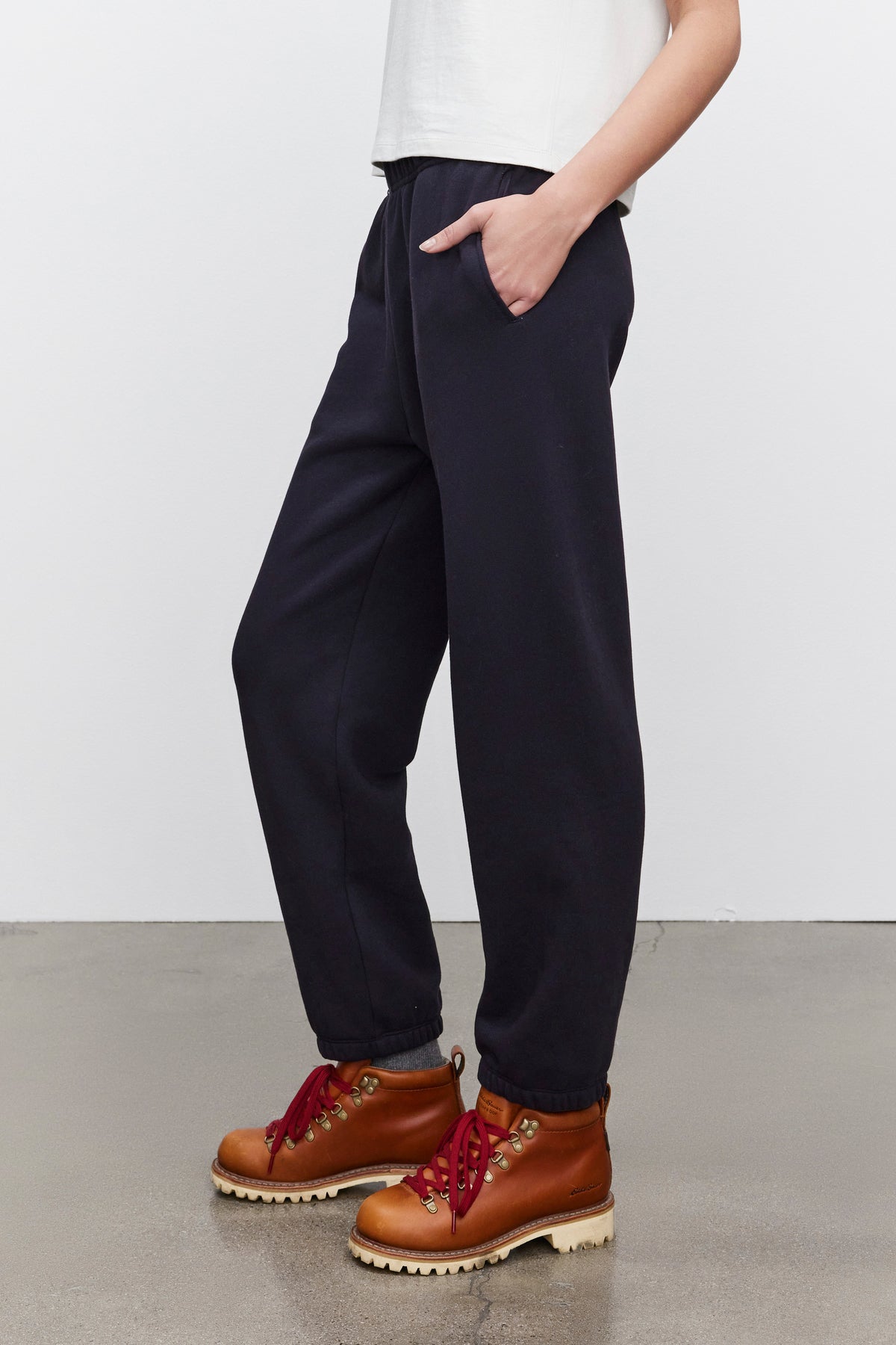   A person dressed in CELESTA SWEATPANTS by Velvet by Graham & Spencer and brown hiking boots with red laces stands against a neutral background, one hand in their pocket, exuding warmth and comfort. 