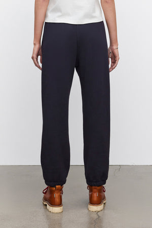 A person is wearing Velvet by Graham & Spencer CELESTA SWEATPANT and brown boots, standing on a concrete floor with their back facing the camera, exuding warmth and comfort.