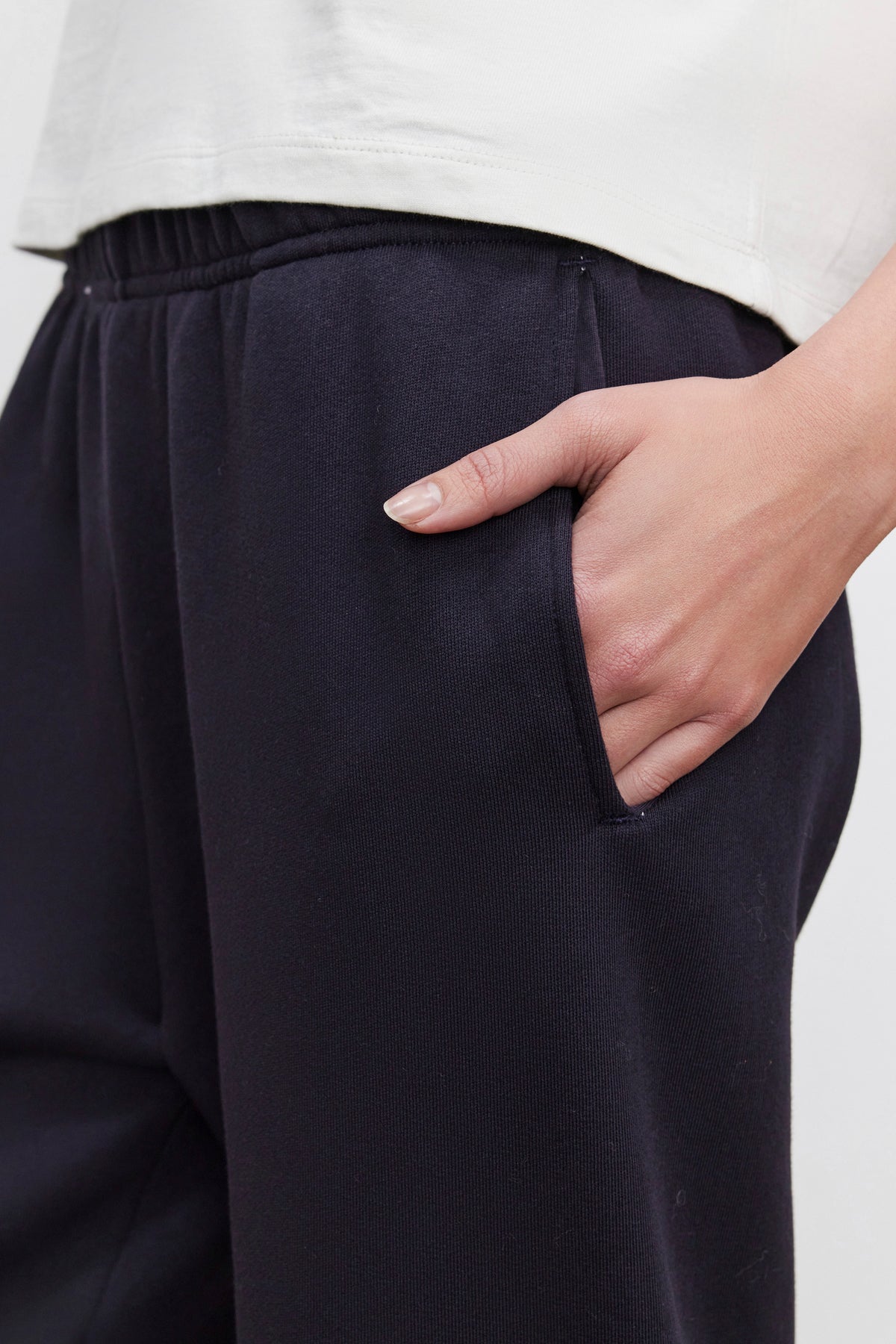   Close-up of a person wearing Velvet by Graham & Spencer's CELESTA SWEATPANT in black cotton fleece, with one hand in their pocket. The warmth and comfort of the pants are complemented by a pristine white shirt. 