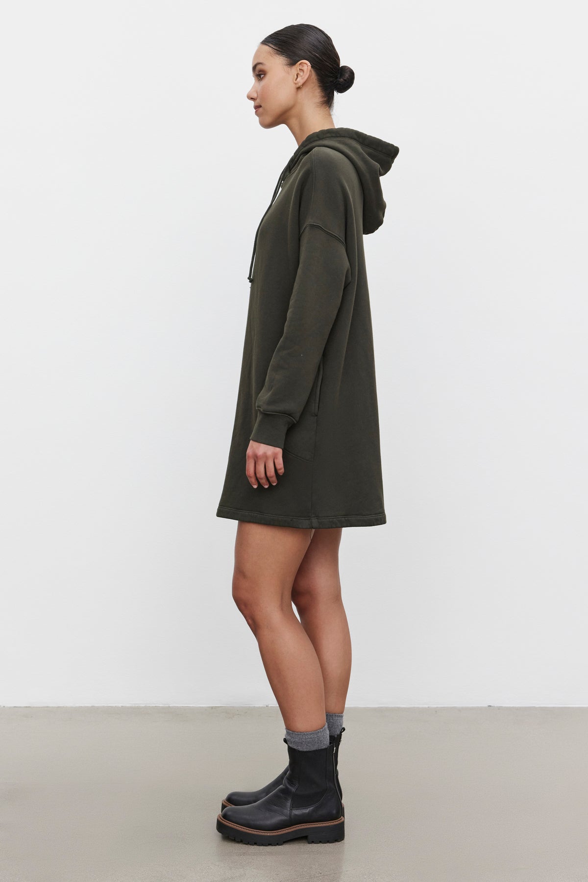   A person stands in profile wearing the ETTA HOODIE DRESS, a dark green, relaxed fit cotton fleece sweatshirt dress by Velvet by Graham & Spencer, paired with black ankle boots and gray socks. The hair is tied back, and the background is plain white. 