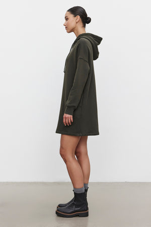 A person stands in profile wearing the ETTA HOODIE DRESS, a dark green, relaxed fit cotton fleece sweatshirt dress by Velvet by Graham & Spencer, paired with black ankle boots and gray socks. The hair is tied back, and the background is plain white.