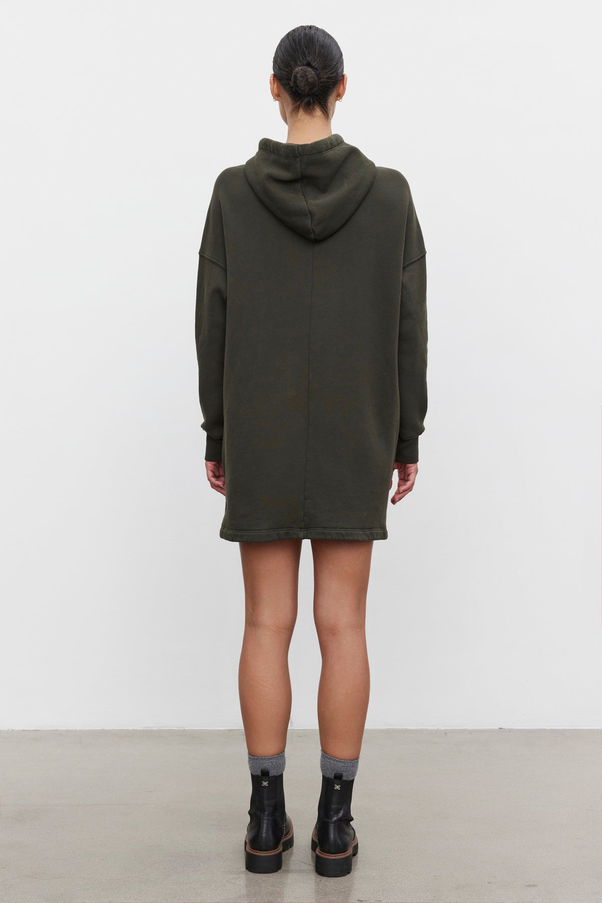   A person with their hair in a bun is standing facing away, wearing a dark green ETTA HOODIE DRESS by Velvet by Graham & Spencer and black ankle boots. 