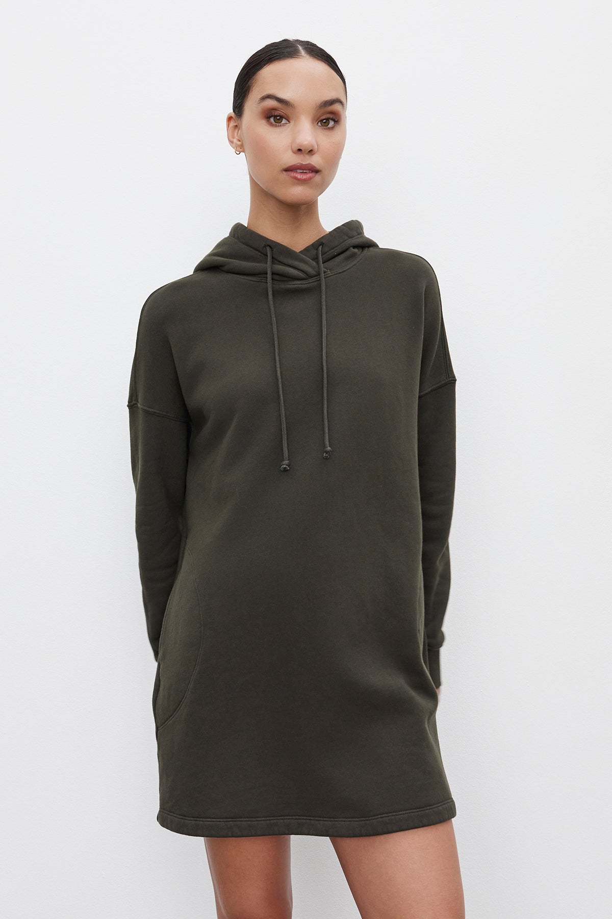   Person standing against a plain white background, wearing the ETTA HOODIE DRESS from Velvet by Graham & Spencer, a relaxed fit, dark green hooded sweatshirt dress made from soft cotton fleece, with their hands behind their back. 