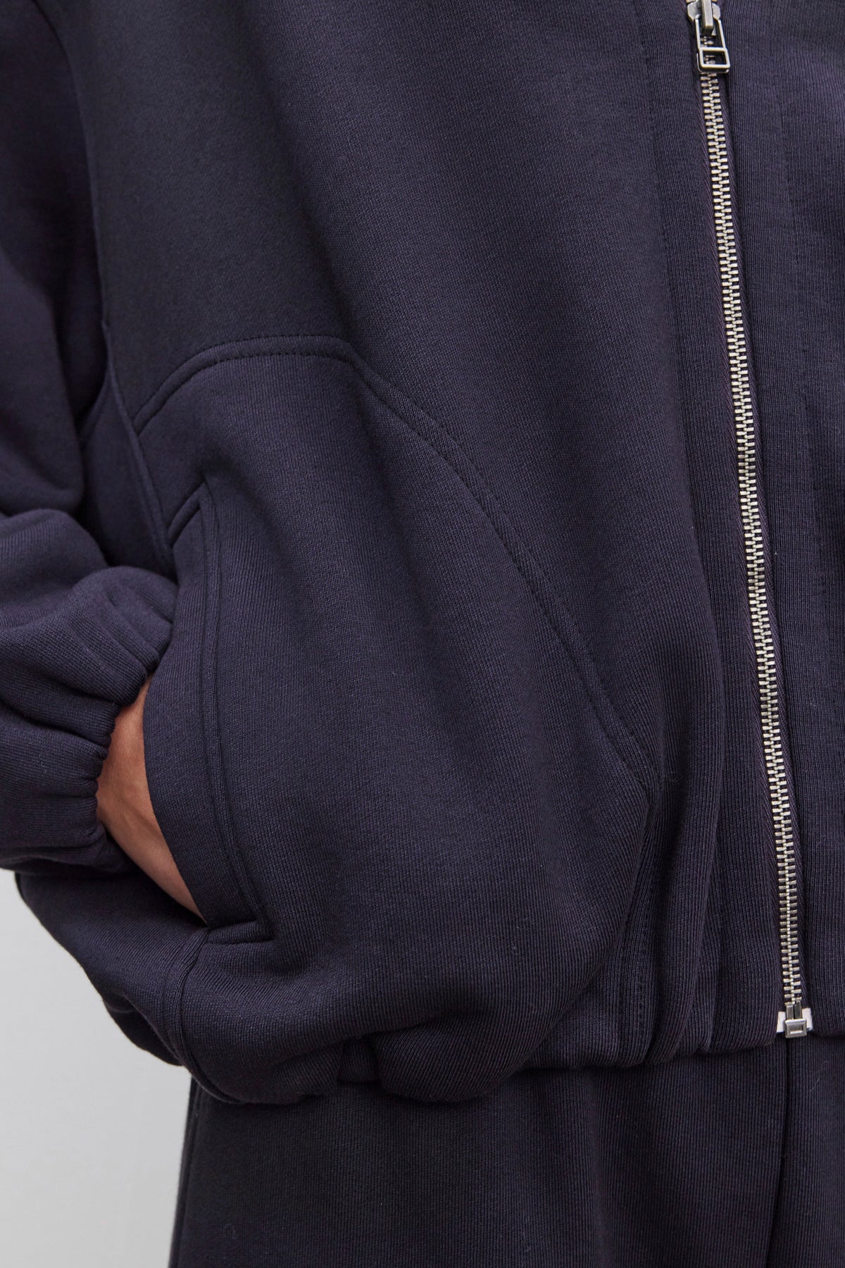 A person wearing an oversized fit, dark GENEVIEVE ZIP-UP SWEATSHIRT by Velvet by Graham & Spencer with their hand in the pocket. The sweatshirt, made from heavier-weight cotton fleece, features a visible zipper and elasticized cuffs.-37856522961089