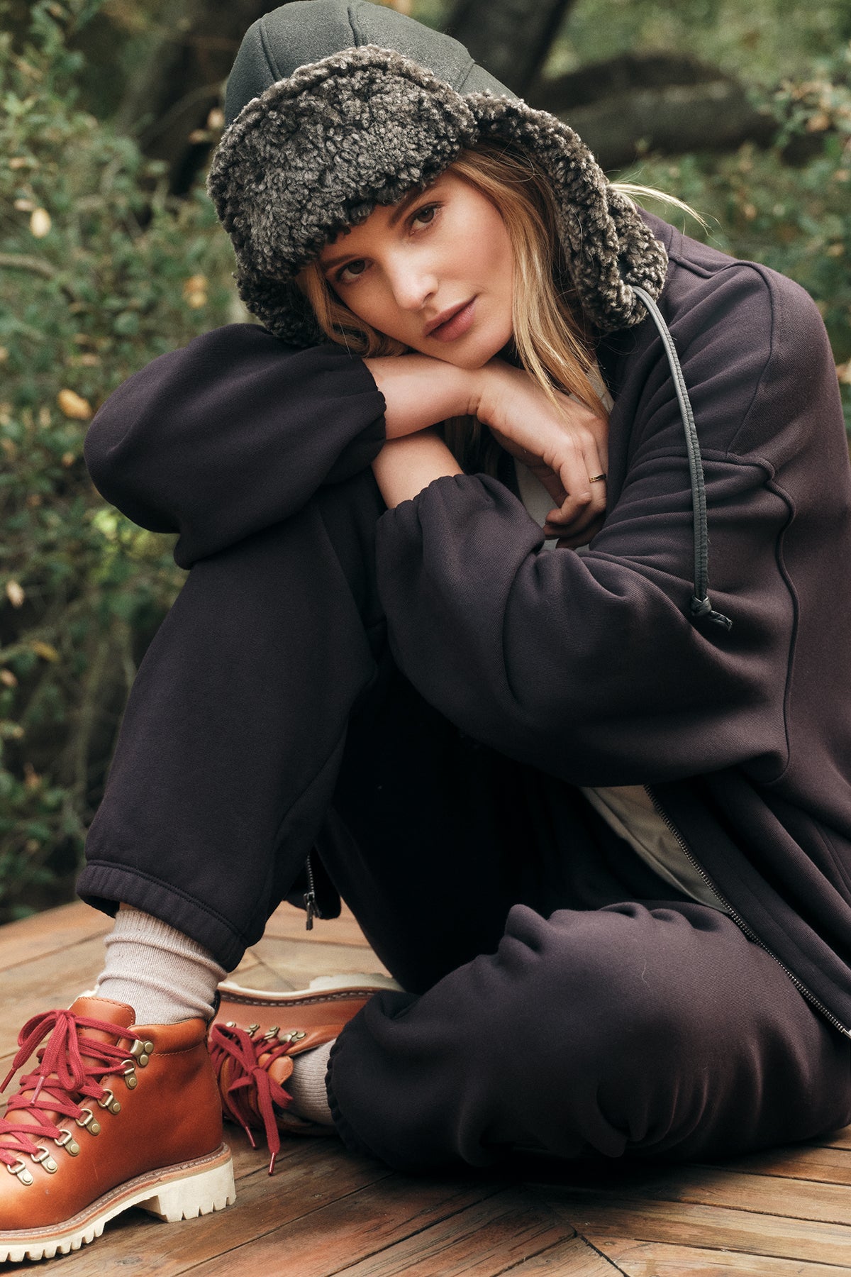 A woman sits outside on a wooden surface, wearing dark sweatpants and the Velvet by Graham & Spencer GENEVIEVE ZIP-UP SWEATSHIRT in an oversized fit, complemented by a wool-lined hat and brown boots with red laces, resting her head on her hand. Green plants are visible behind her.-37856523059393