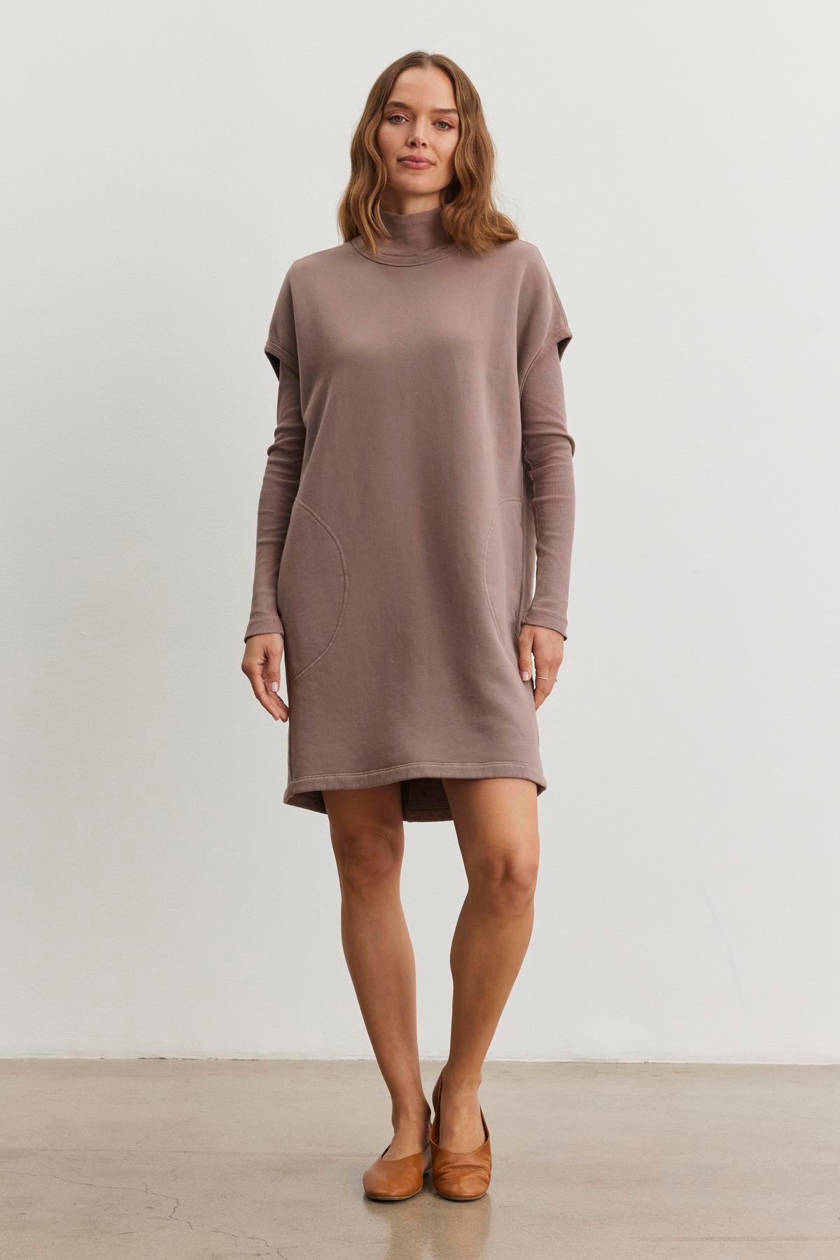 A woman wearing the LITTA DRESS by Velvet by Graham & Spencer is standing on a light-colored floor against a plain white background, showcasing its taupe turtleneck design with pockets and sweatshirt style.-38483614302401