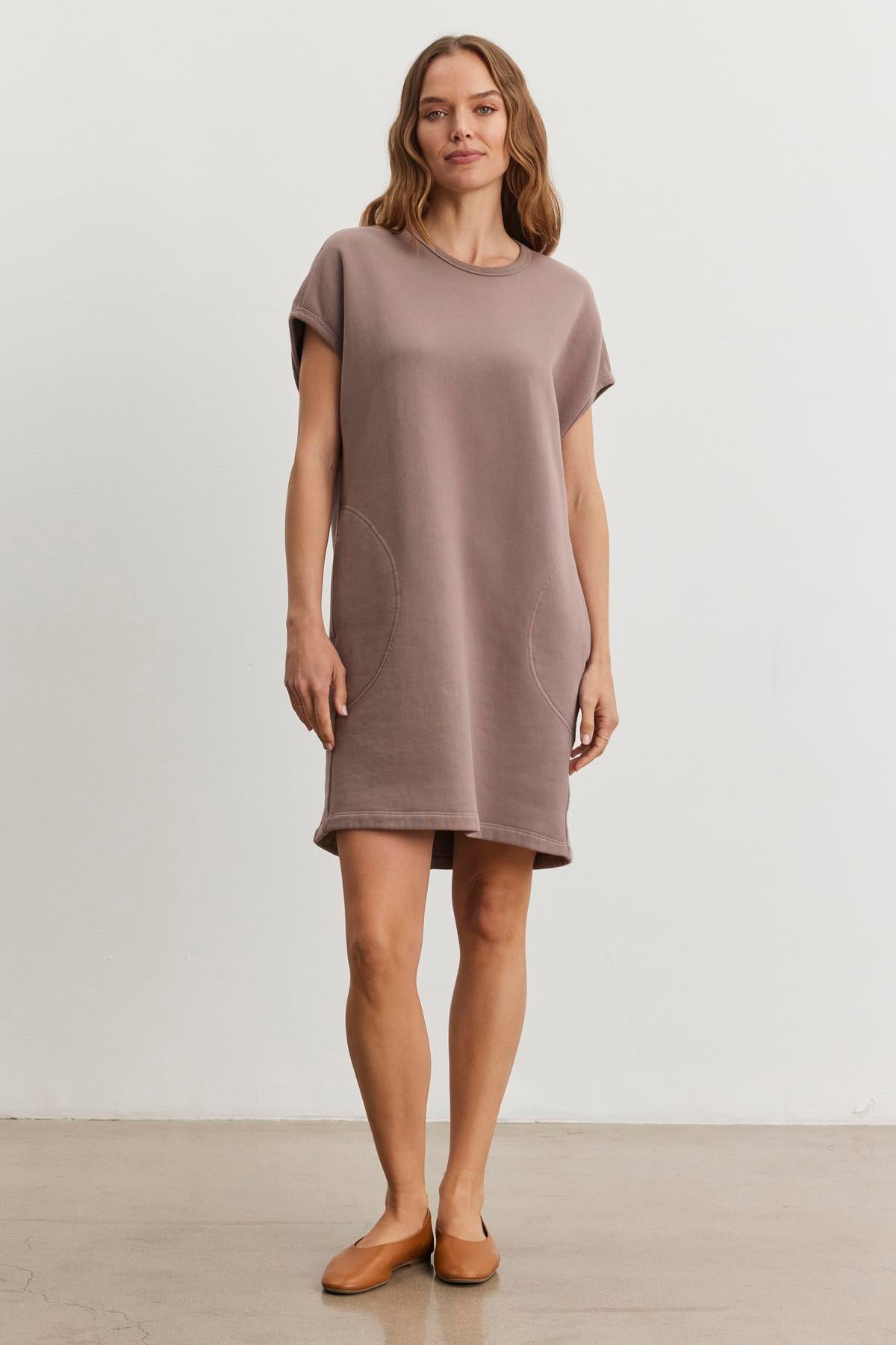 A woman exudes effortless style on a polished concrete floor, dressed in the LITTA DRESS by Velvet by Graham & Spencer, featuring a loose fit, short sleeves, pockets, and a subtle sweatshirt style in taupe.-38483614400705