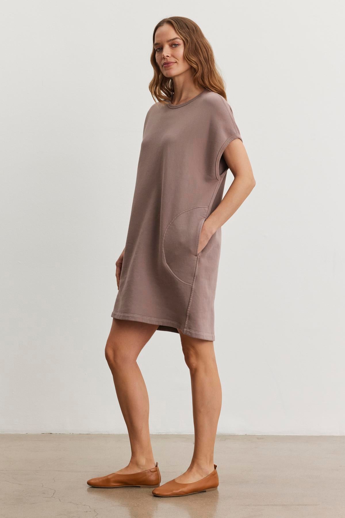 The woman stands on a concrete floor against a plain white wall, wearing the LITTA DRESS by Velvet by Graham & Spencer, a loose brown t-shirt dress featuring side pockets. Its sweatshirt style and crew neckline enhance the comfort, complemented by her tan flat shoes.-38483614367937