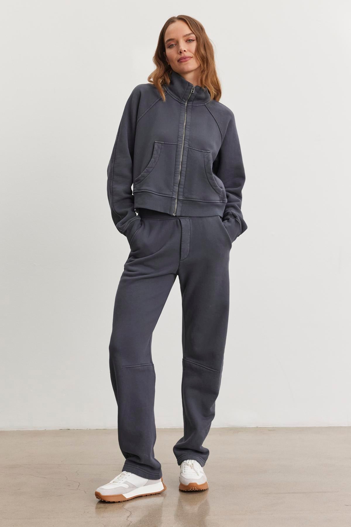   Wearing the MATTI SWEATPANT by Velvet by Graham & Spencer, a person stands confidently in a cozy dark gray tracksuit. The fit features an elastic waist and hands tucked into pockets, complemented by white sneakers, all set against a plain background with a neutral expression. 