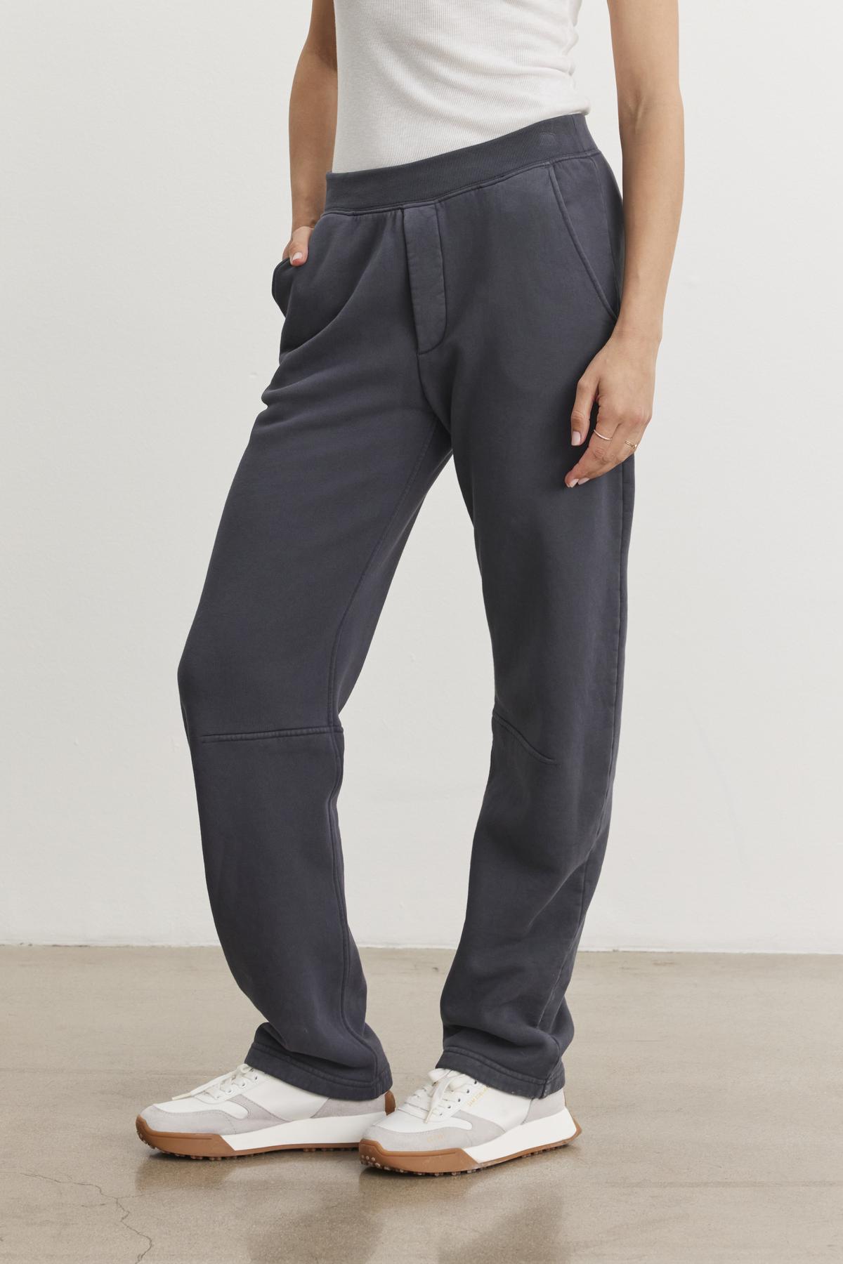 A person wearing a white shirt, MATTI SWEATPANT from Velvet by Graham & Spencer in cozy dark gray with an elastic waist, and white sneakers stands with hands in pockets on a light-colored floor.-38483616235713