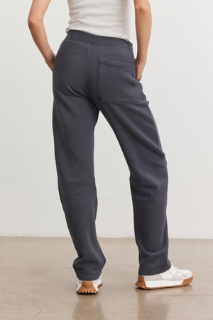 A person wearing a white tank top and the MATTI SWEATPANT from Velvet by Graham & Spencer in dark gray, featuring an elastic waist, stands on a concrete floor with their back to the camera, hands on hips, and white shoes with brown soles.