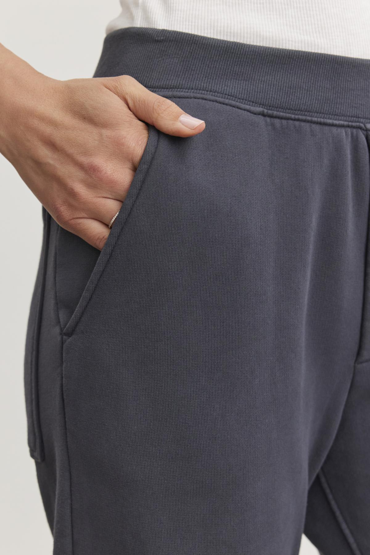   Close-up of a person wearing dark gray MATTI SWEATPANT from Velvet by Graham & Spencer, with one hand in the pocket, showcasing their comfortable fit and elastic waist. 