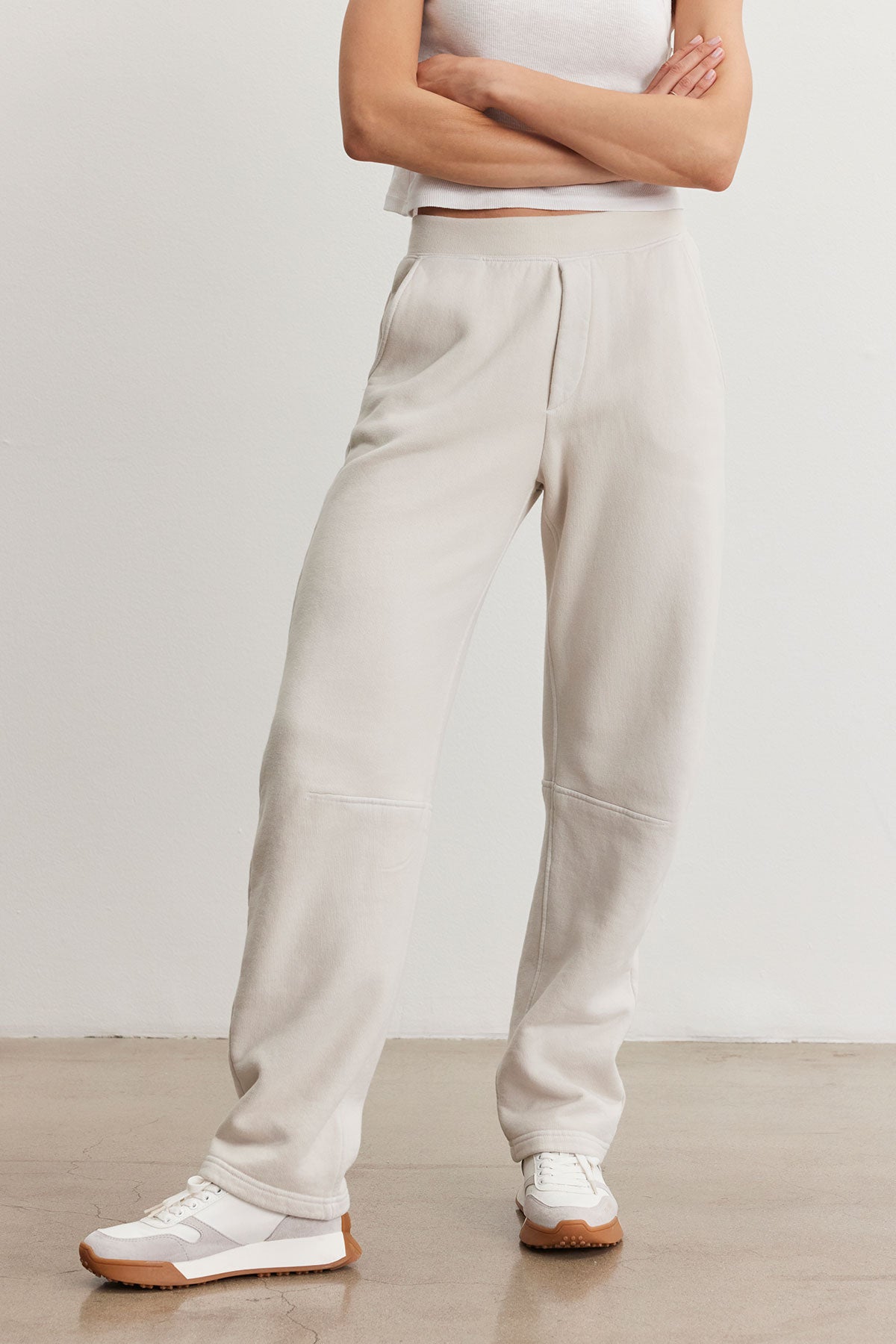 A person wearing a white sleeveless top, the MATTI SWEATPANT by Velvet by Graham & Spencer in light beige cotton fleece, and white sneakers stands with arms crossed against a plain background, effortlessly showcasing a laid-back style and comfortable fit.-38301388308673