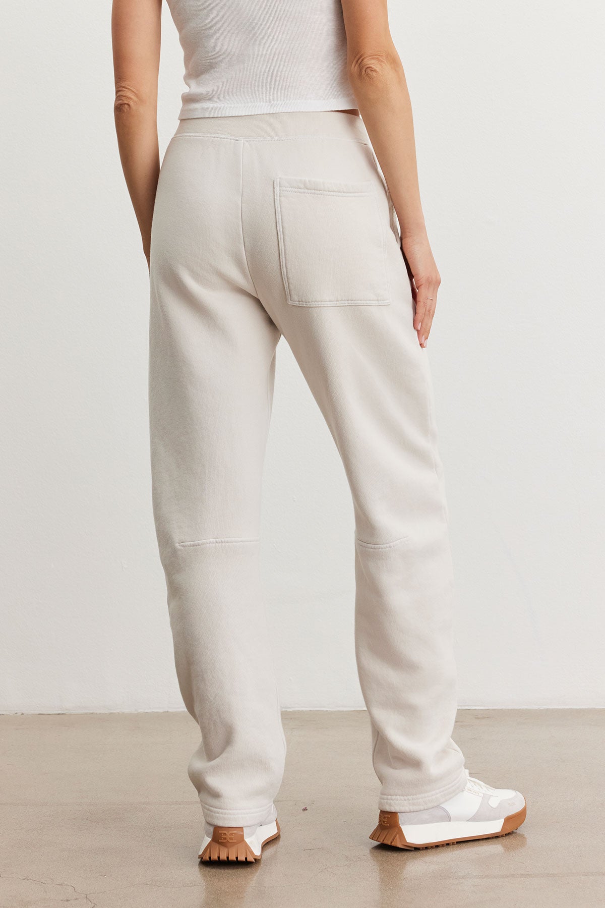   A person embodies a relaxed style, wearing the light beige MATTI SWEATPANT from Velvet by Graham & Spencer along with a white top, standing on a light brown floor and facing away from the camera. White sneakers perfectly complete this comfortable ensemble. 