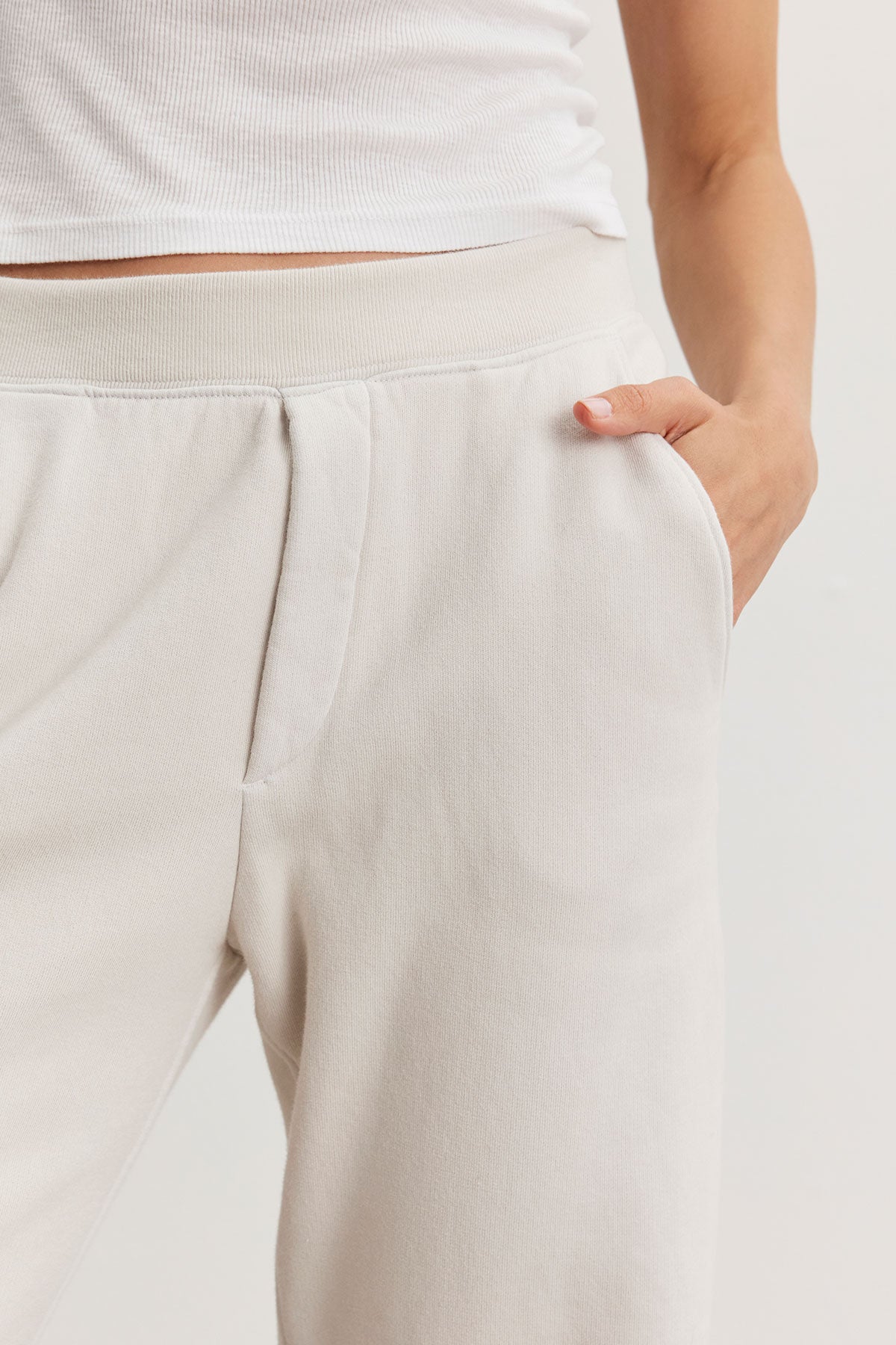   A person exudes a laid-back style, donning a white tank top paired with the light beige MATTI SWEATPANT from Velvet by Graham & Spencer, which offers a comfortable fit, one hand casually tucked in the pocket. 