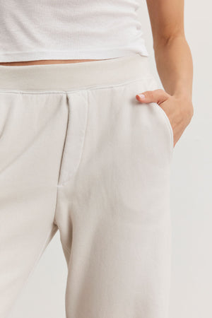 A person exudes a laid-back style, donning a white tank top paired with the light beige MATTI SWEATPANT from Velvet by Graham & Spencer, which offers a comfortable fit, one hand casually tucked in the pocket.