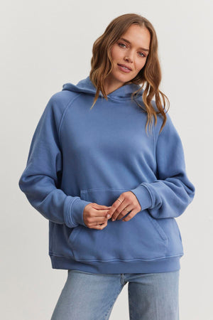 A person wearing the oversized VERONICA HOODIE in blue, by Velvet by Graham & Spencer, along with jeans, stands against a plain backdrop.