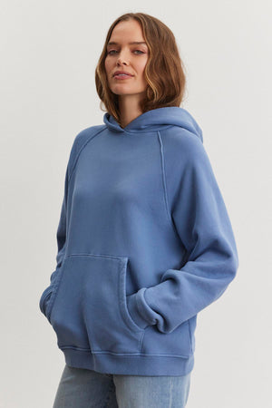 A person wearing the oversized VERONICA HOODIE by Velvet by Graham & Spencer, crafted from cotton fleece with hands snugly tucked in its front pocket, stands against a plain background.