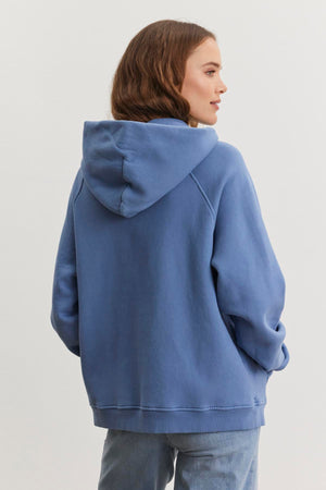 A person wearing a Velvet by Graham & Spencer's oversized VERONICA HOODIE in blue cotton fleece and jeans is standing with their back to the camera.