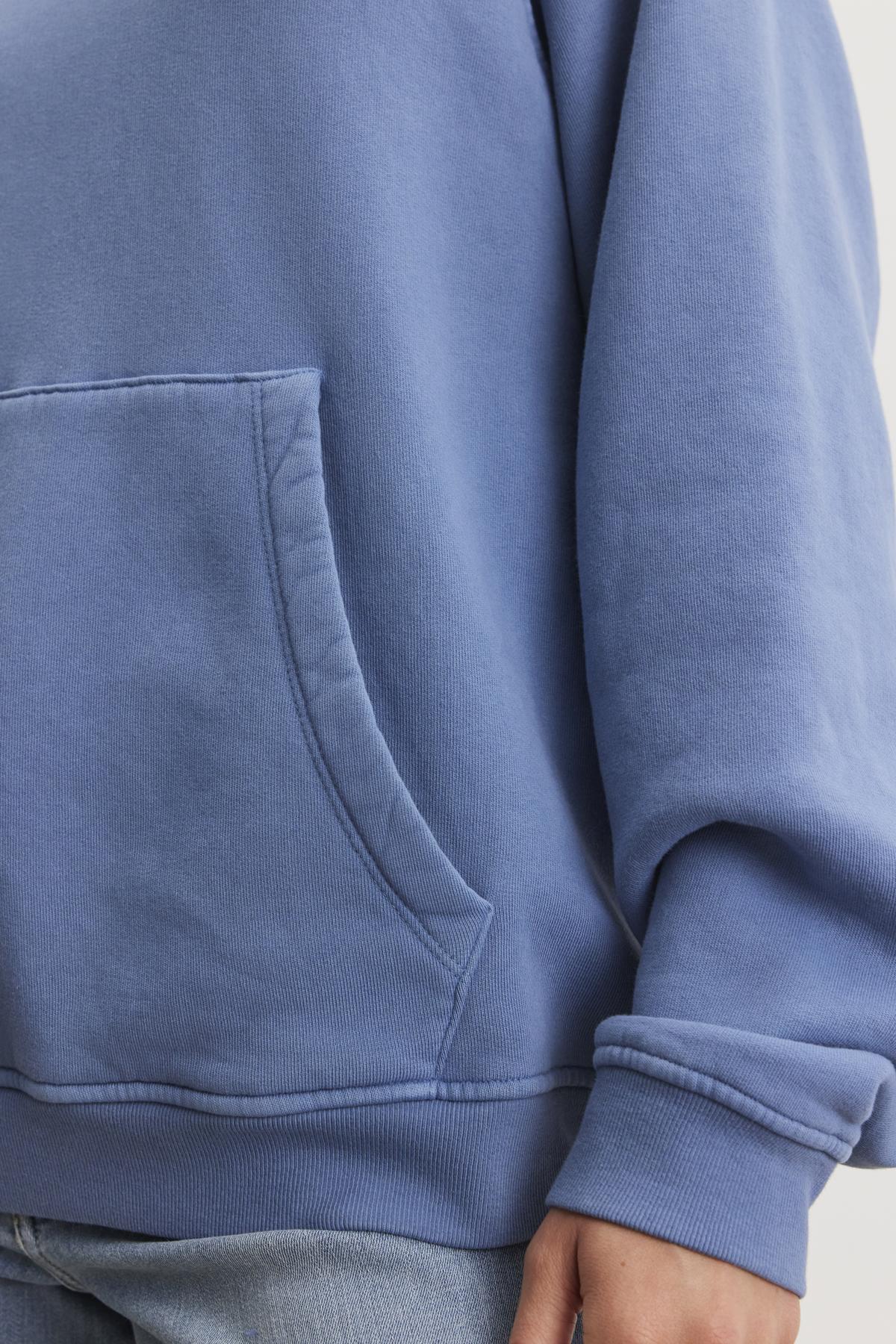 Close-up of a person wearing the VERONICA HOODIE by Velvet by Graham & Spencer, featuring a blue cotton fleece pullover style with a front pocket, paired with light blue jeans.-38512021766337