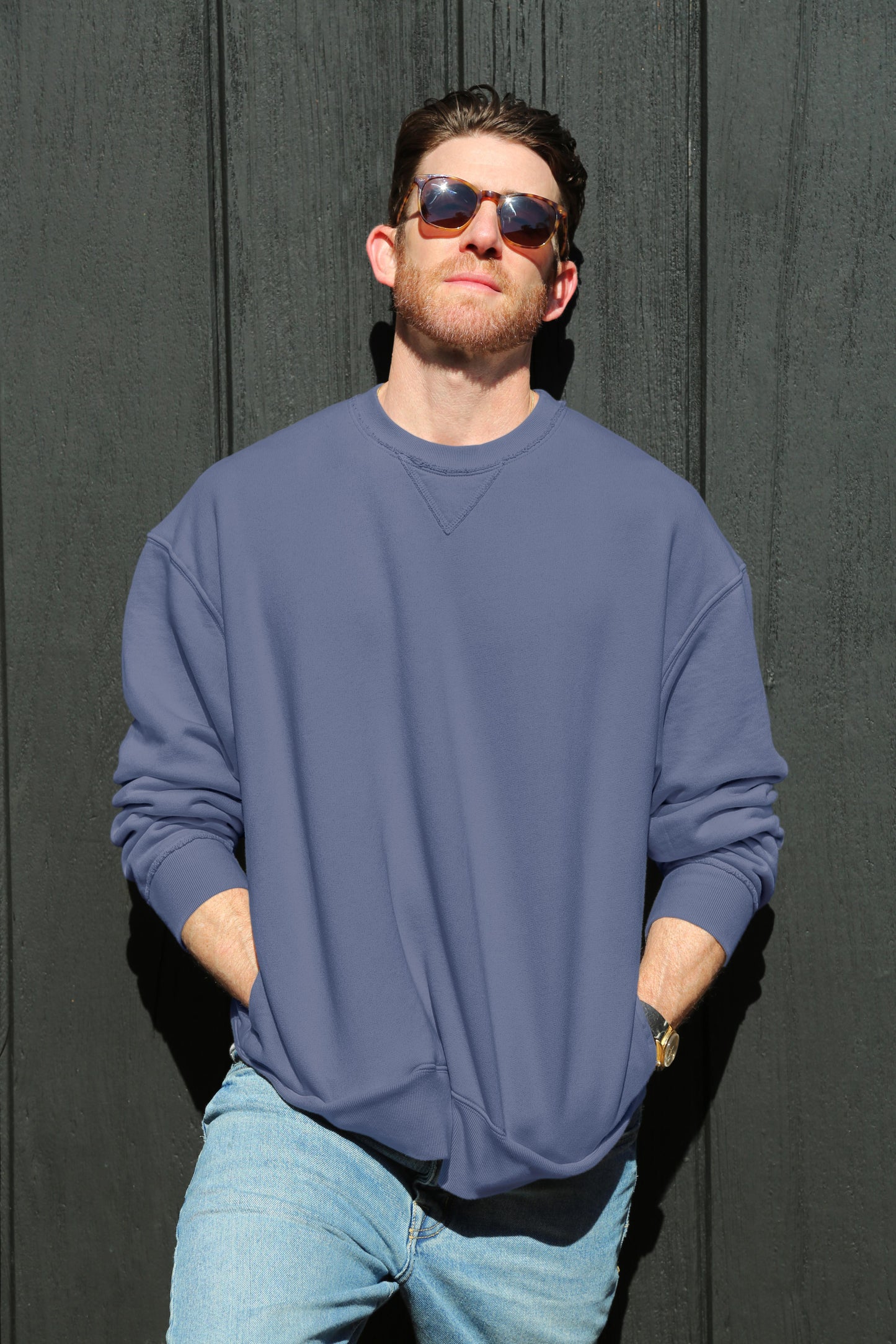 A man in sunglasses stands with hands in his pockets, wearing the Velvet by Graham & Spencer FORREST SWEATSHIRT, a relaxed fit cotton fleece, paired with jeans and leaning against a dark wall.-38325785428161