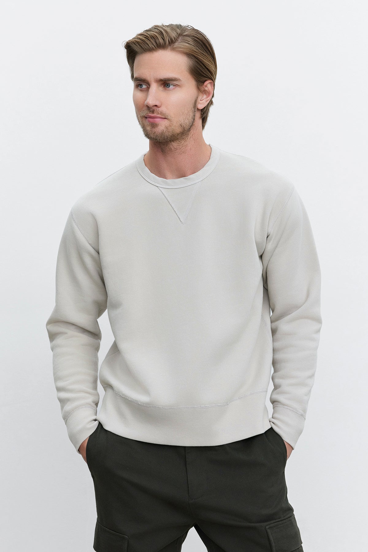   A man wearing the ISAMU SWEATSHIRT by Velvet by Graham & Spencer, characterized by its light gray vintage look, and dark pants stands against a plain white background with hands in pockets. 