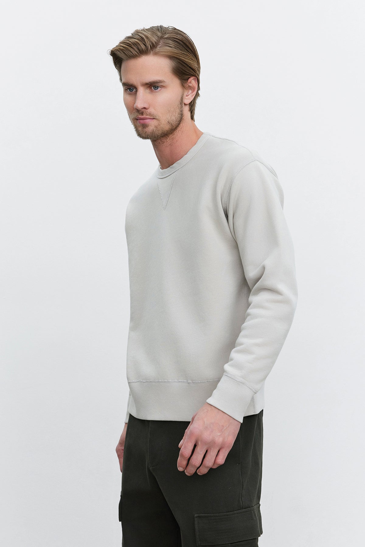   A man wearing the ISAMU SWEATSHIRT by Velvet by Graham & Spencer, paired with dark pants, stands against a plain white background. He has short hair and a beard, and he gazes to the side. 