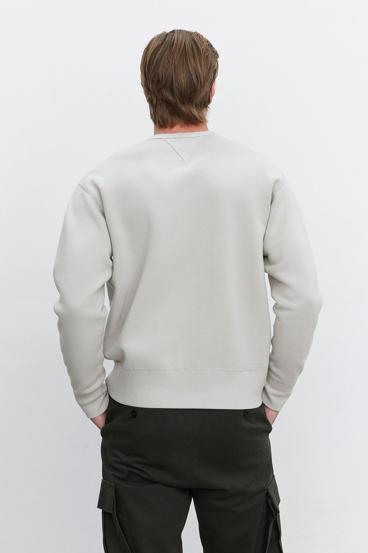   A person with light brown hair is standing with their back to the camera, wearing a light gray ISAMU SWEATSHIRT from Velvet by Graham & Spencer and dark green pants, creating a timeless appearance against the plain white background. 