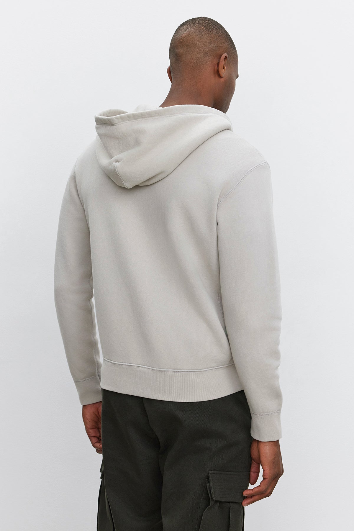 A person stands facing away, wearing a light gray REN HOODIE by Velvet by Graham & Spencer and dark cargo pants against a plain background, giving off a vintage look.-37893921112257