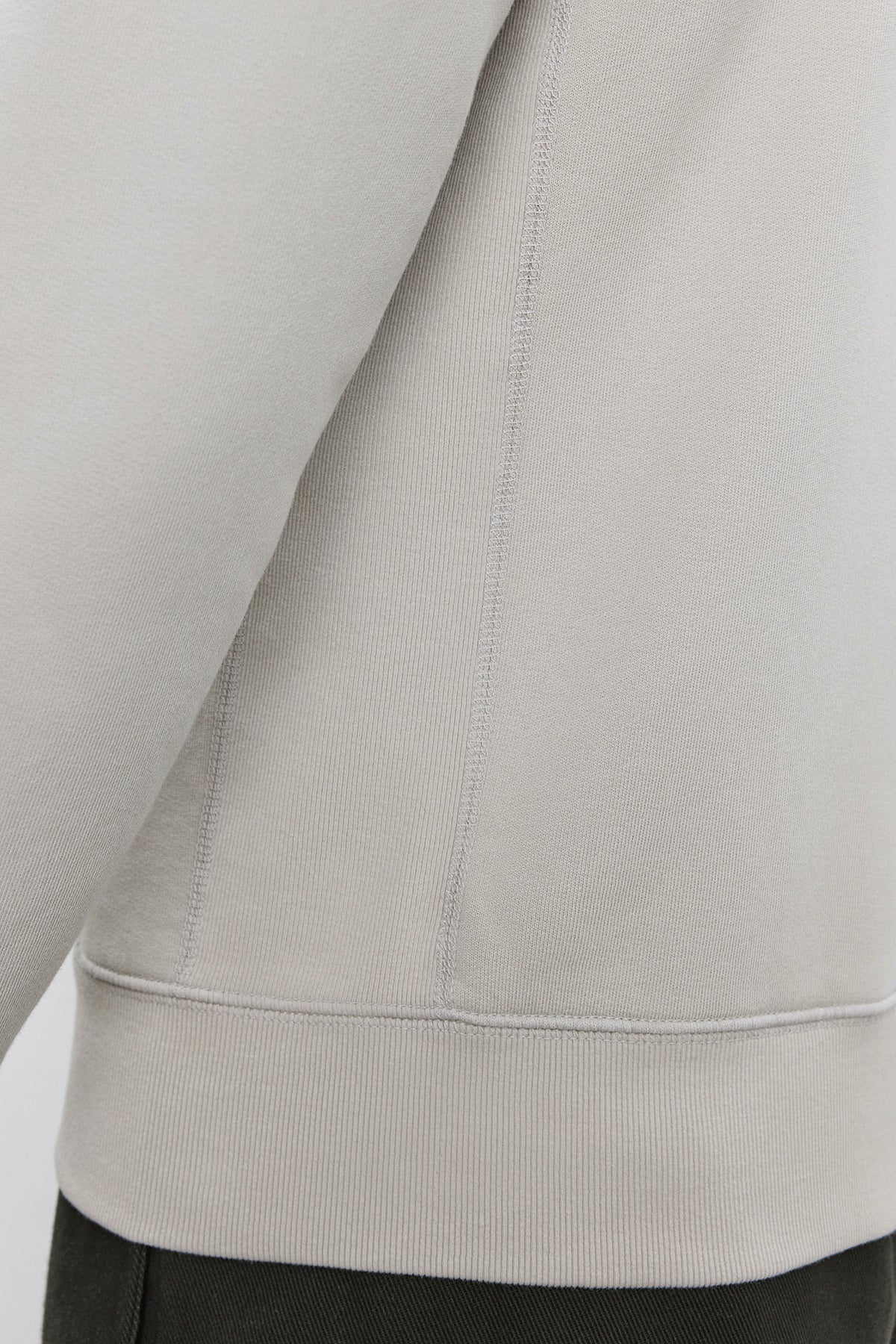   Close-up of the side seam and lower part of a light beige REN HOODIE by Velvet by Graham & Spencer, made from cotton fleece, with the hemline visible, worn over dark green pants. 