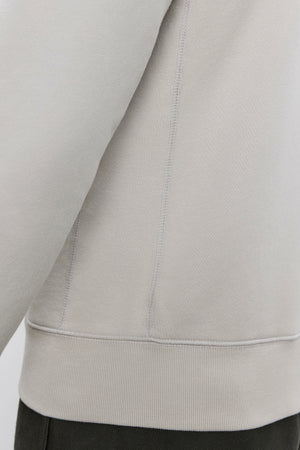 Close-up of the side seam and lower part of a light beige REN HOODIE by Velvet by Graham & Spencer, made from cotton fleece, with the hemline visible, worn over dark green pants.