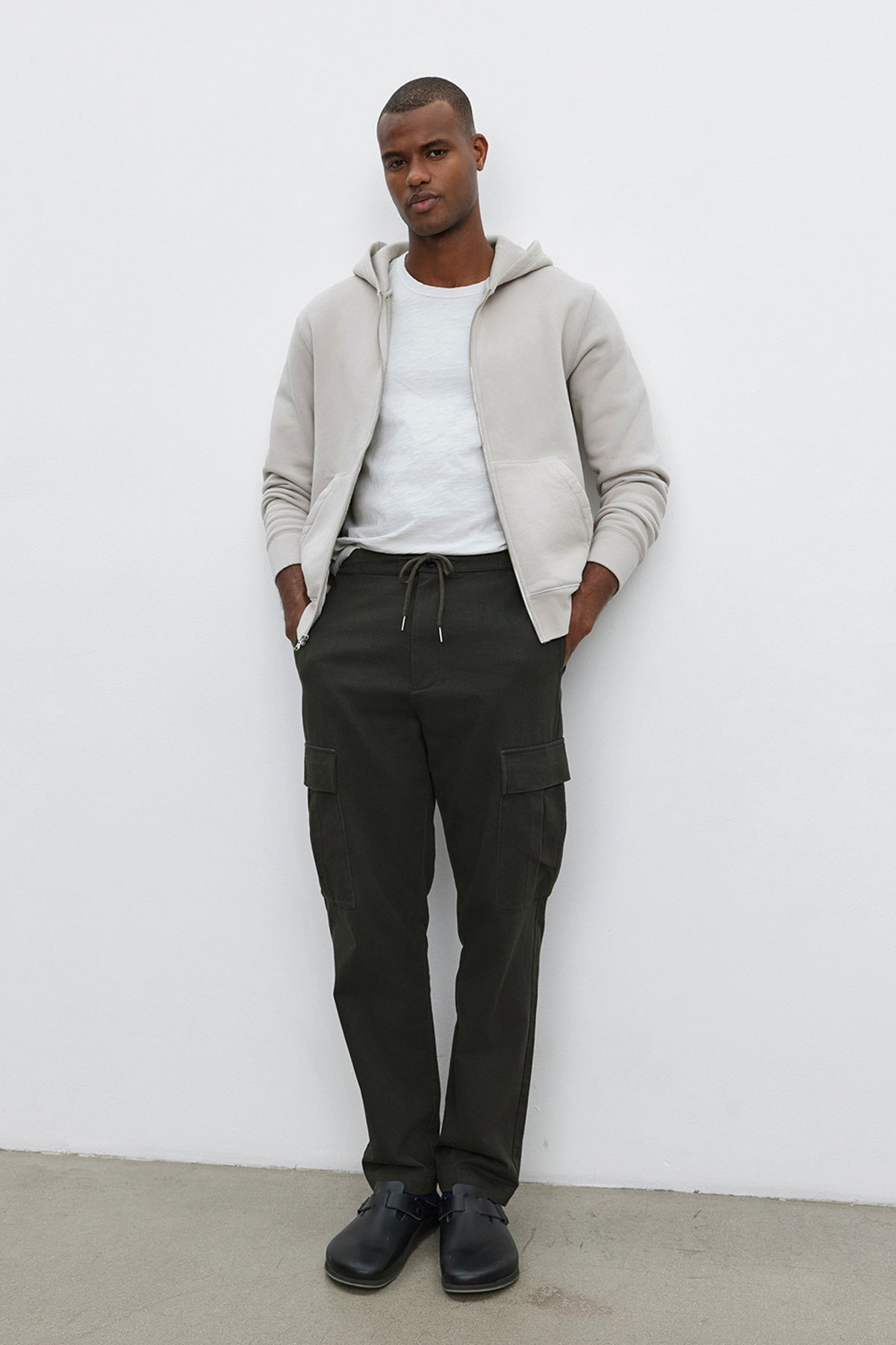   A man stands against a white wall wearing the REN HOODIE by Velvet by Graham & Spencer, a white t-shirt, dark green cargo pants, and black shoes, with his hands in his pockets. 